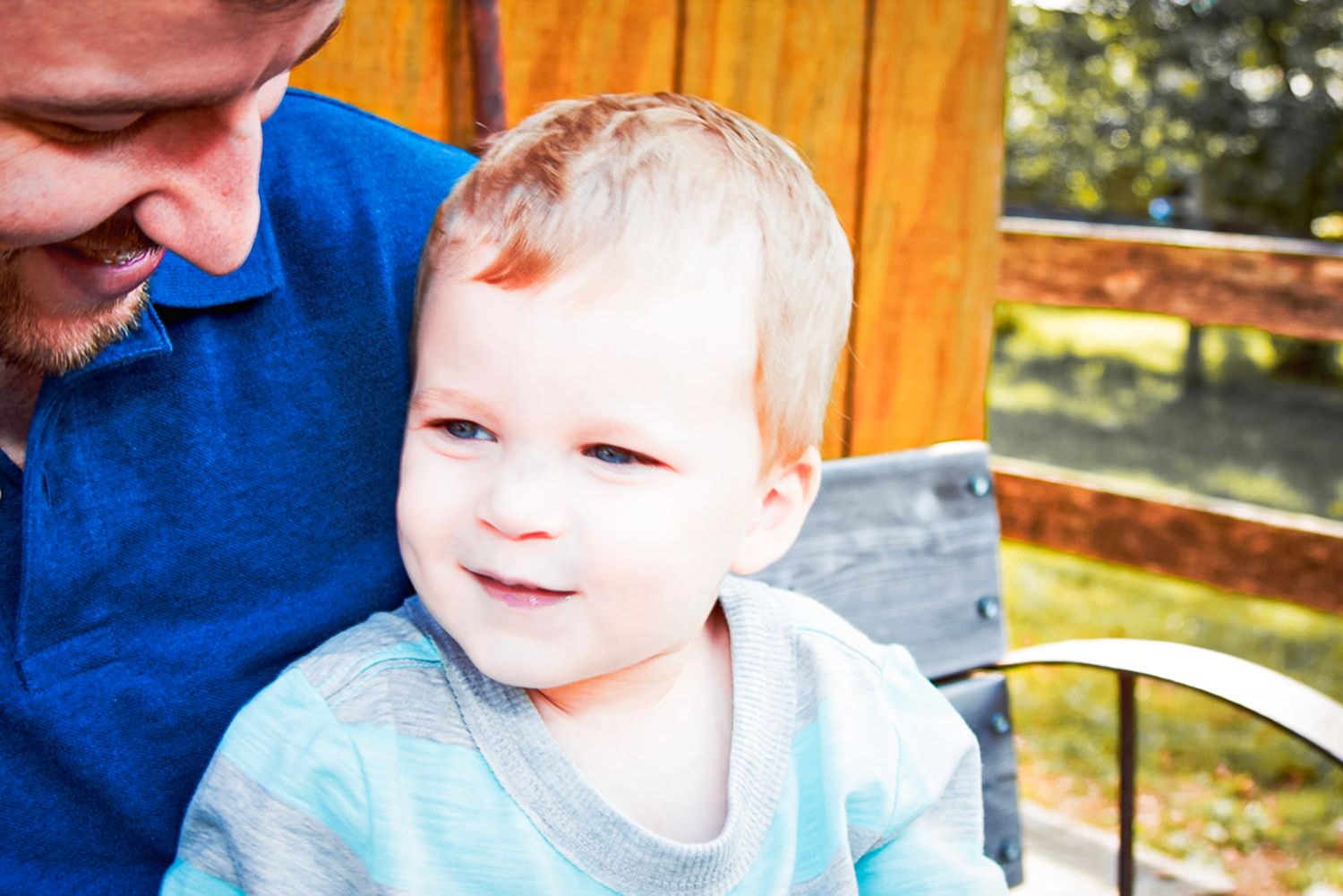A smiling toddler with blonde hair sits with an adult in a blue shirt outdoors on a sunny day.