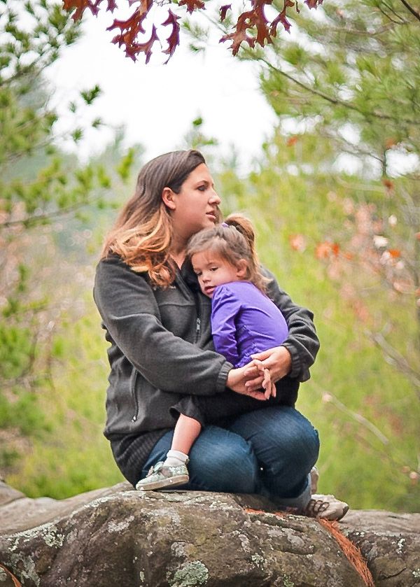 A woman sits on a rock outdoors, cradling a young child in a purple shirt, surrounded by autumn foliage.