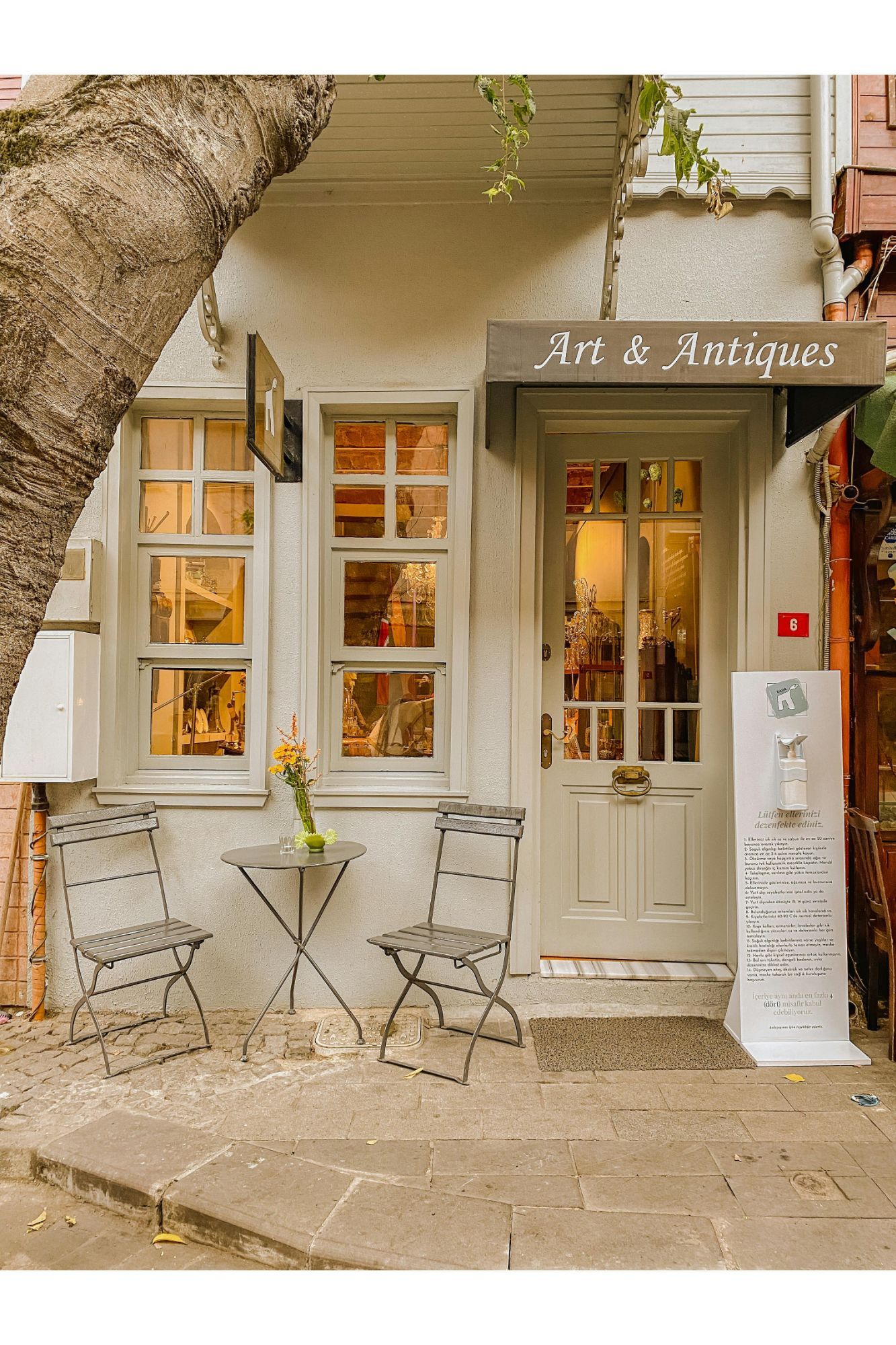 Charming Art & Antiques shop exterior with bistro chairs, warm glowing windows, and a welcoming door on a quaint cobblestone street.