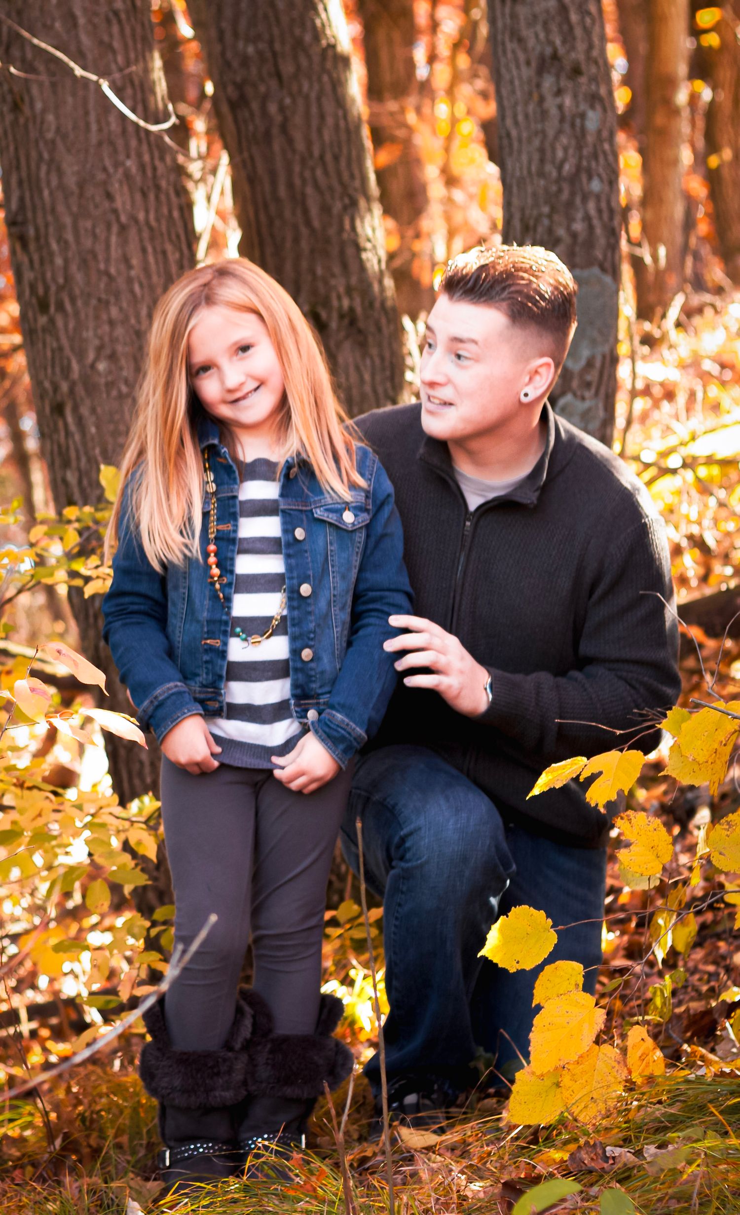 A parent and young blonde girl pose together in a colorful autumn forest, wearing casual jackets and smiling warmly.