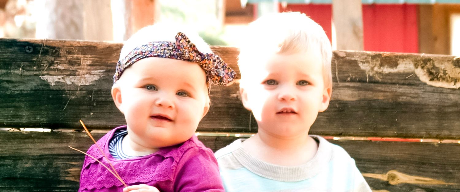 Two adorable baby girls sitting together outdoors, one wearing a purple outfit with a bow headband.