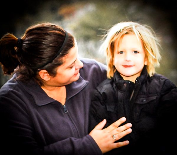 A woman smiles at a young blonde child in a black jacket, sharing a warm moment outdoors in natural light.