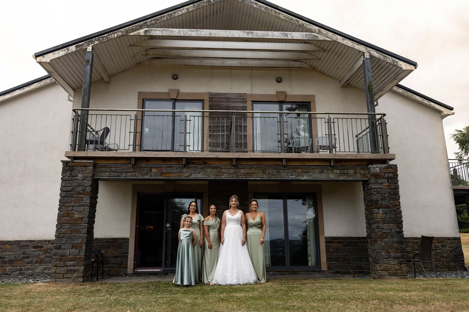 Bride in white gown poses with three bridesmaids in sage green dresses outside a stone wedding venue.