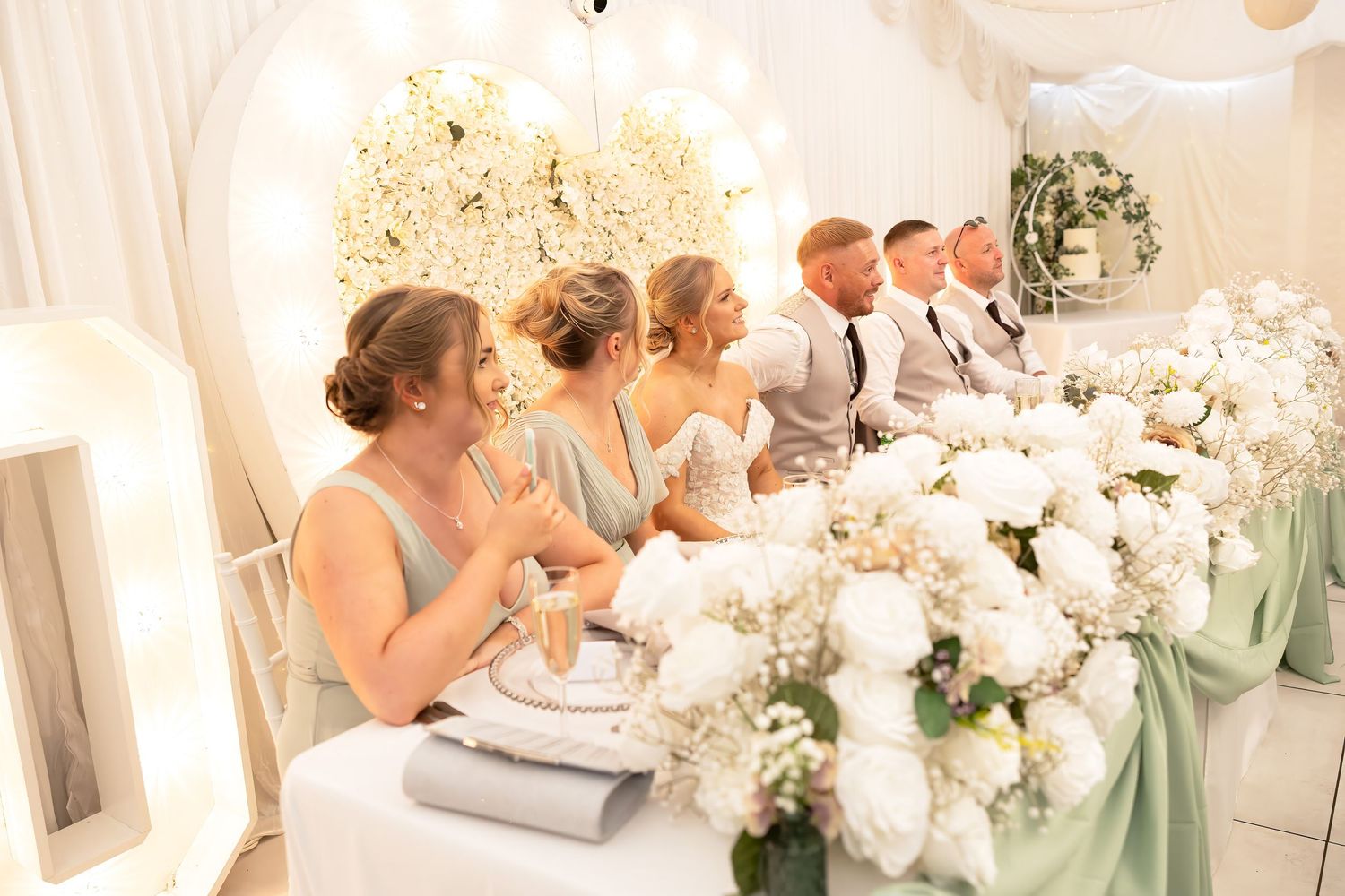 Wedding party seated at head table with white hydrangea centerpieces and glowing floral backdrop.