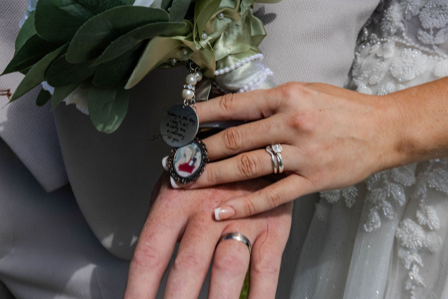 Close-up of bride and grooms hands showing wedding rings and memorial locket on bouquet.