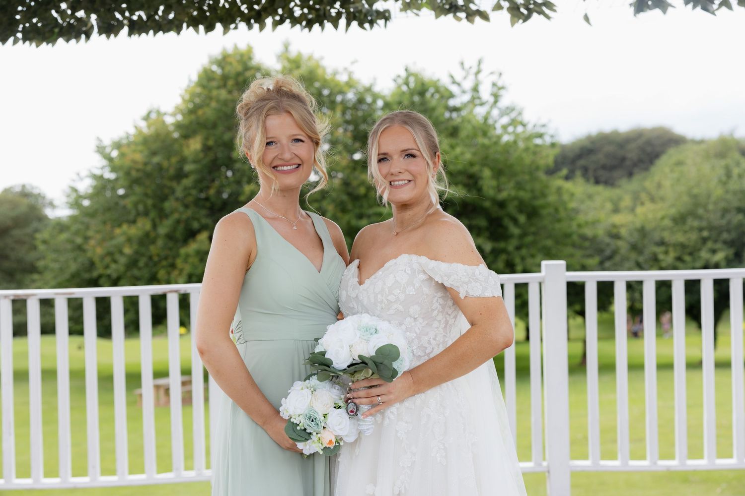 Bride in white off-shoulder gown holding bouquet posing with bridesmaid in sage green dress outdoors.