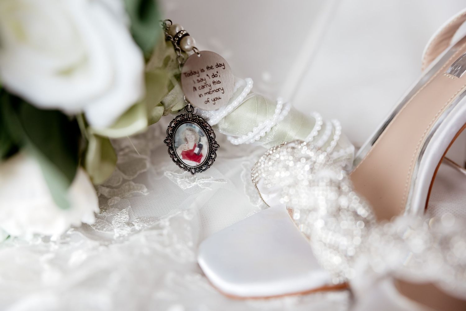 Close-up of bridal accessories including a memorial locket charm, pearl bracelet, and white satin wedding shoes.