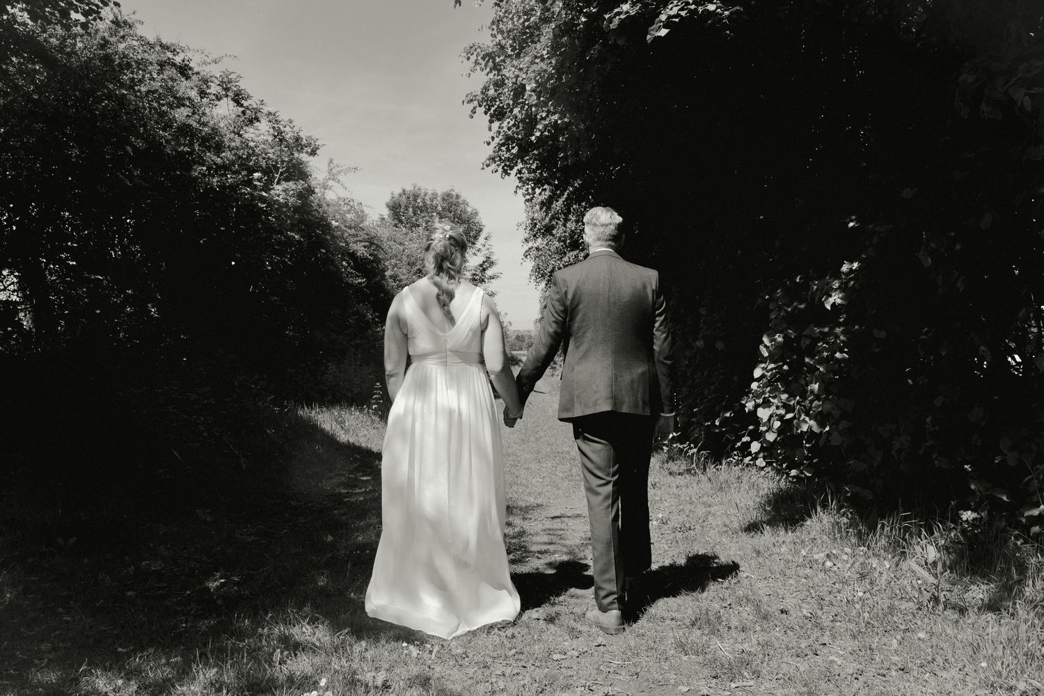 Black and white photo of bride and groom walking hand in hand down a tree-lined path.
