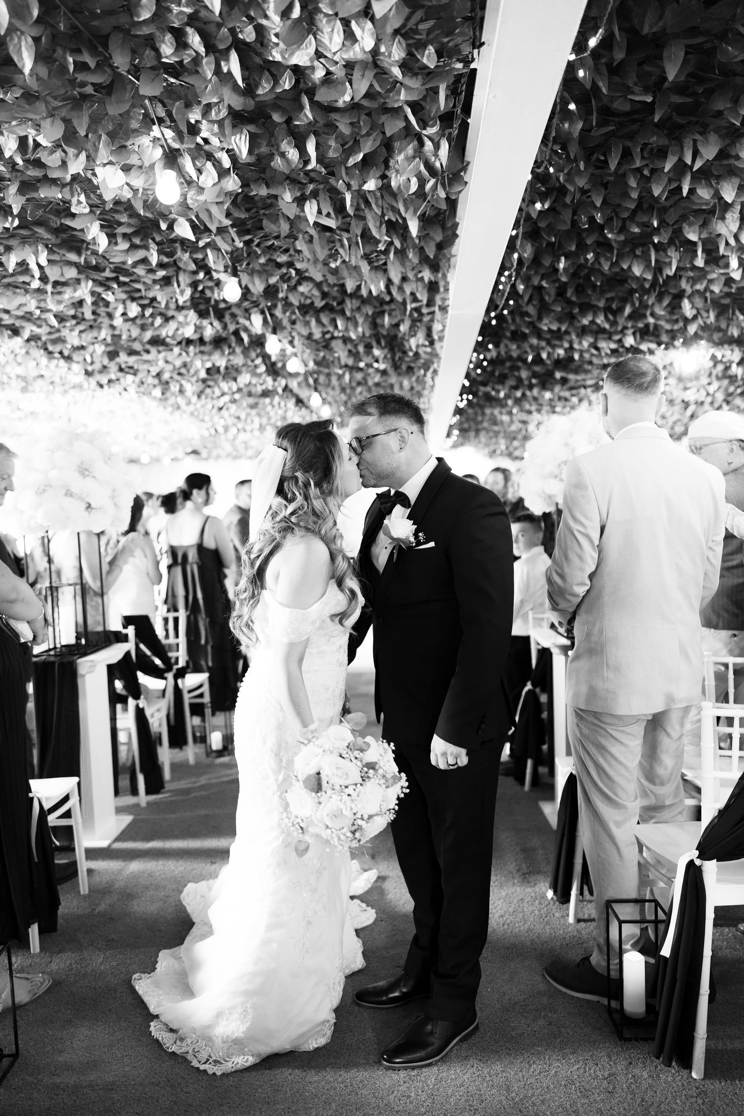 Black and white photo of bride and groom kissing under stunning leafy ceiling at wedding reception.