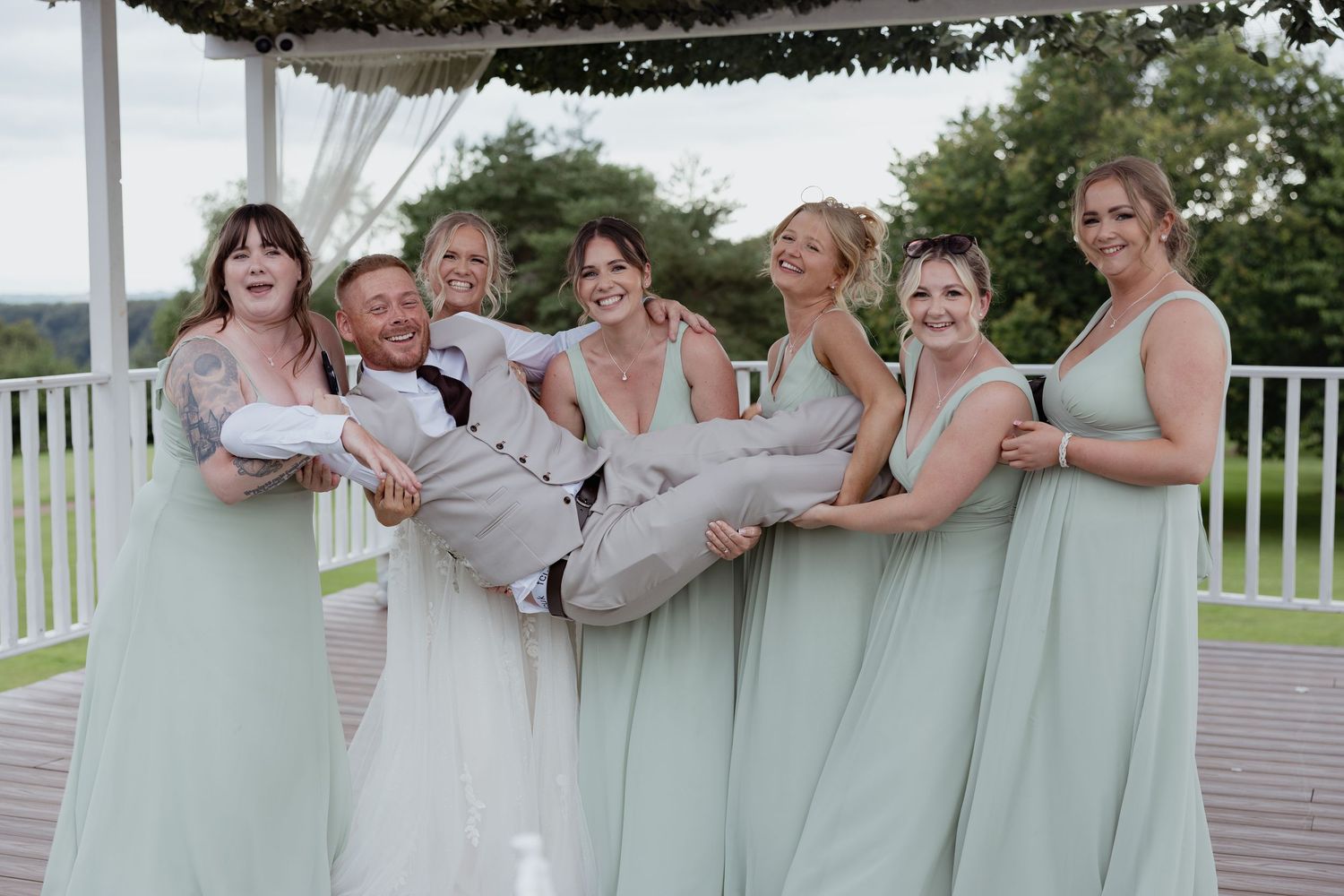 Bridesmaids in sage green dresses playfully lifting the groom while the bride laughs on a deck.