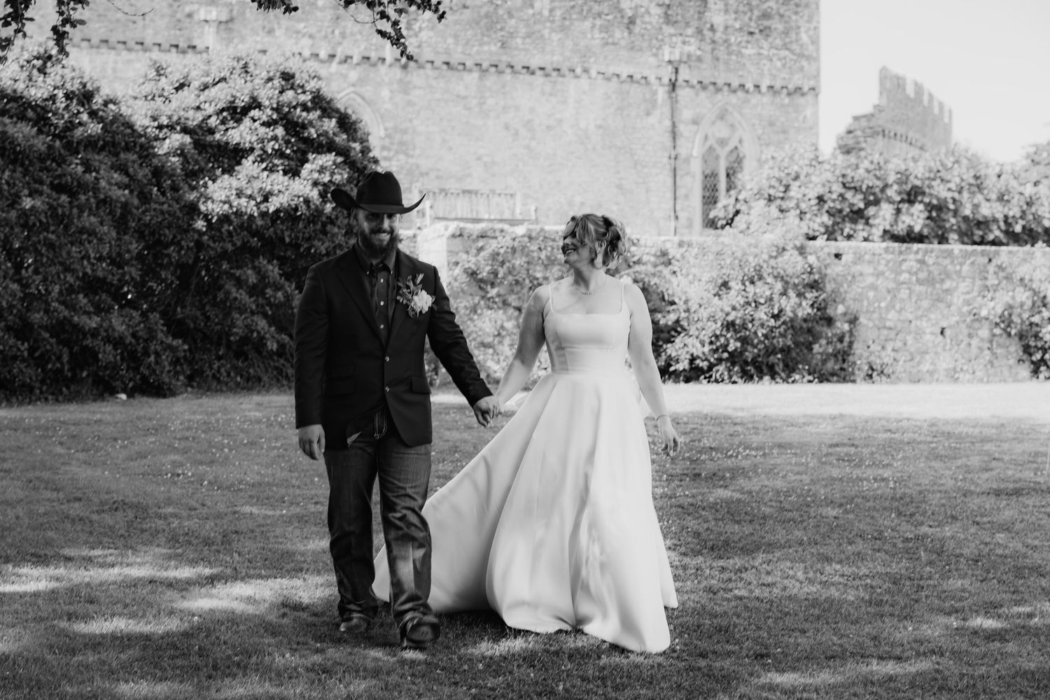Black and white photo of bride and groom walking hand in hand on grass near historic castle.