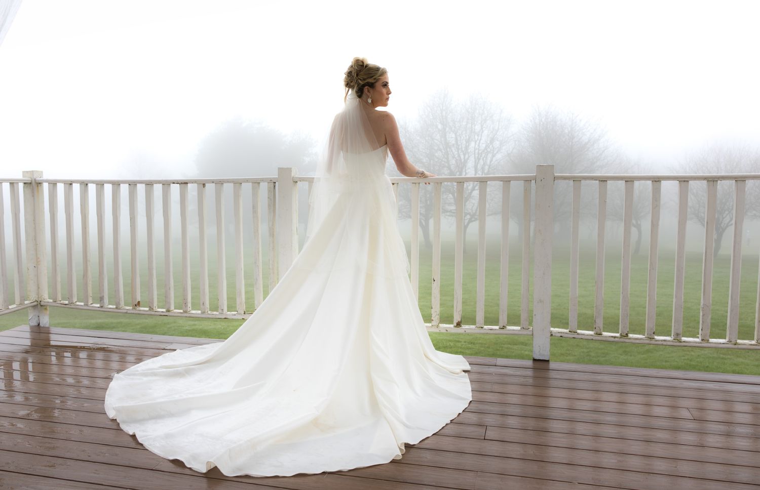 Bride in elegant white wedding gown with long train standing on deck overlooking misty landscape.