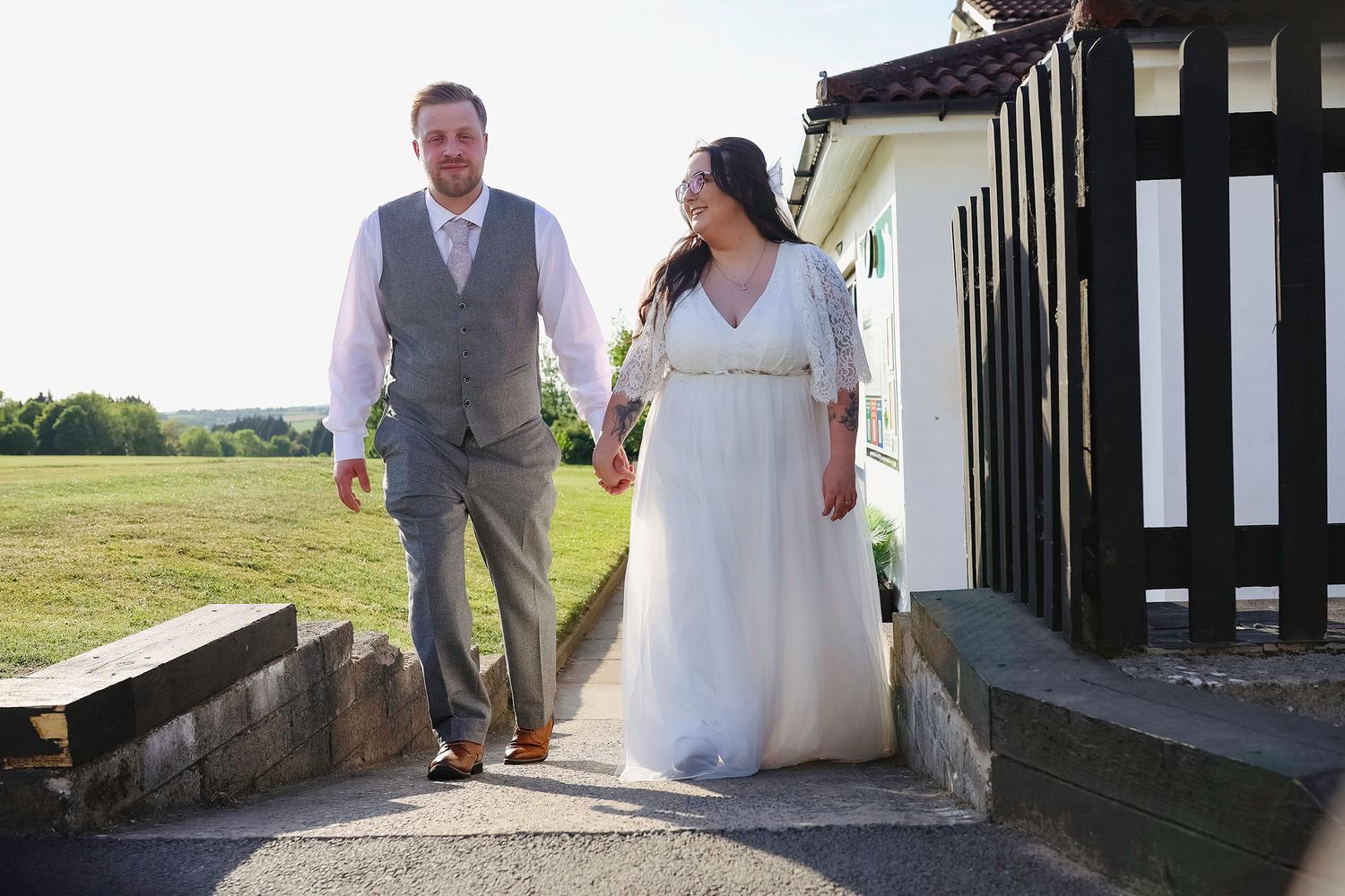 Bride in white gown and groom in grey suit hold hands walking outdoors on sunny wedding day. South Wales weddings