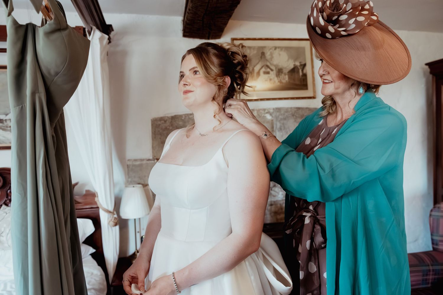 A bride in a white gown gets her necklace fastened by a woman in a teal dress and brown hat in a cozy room.
