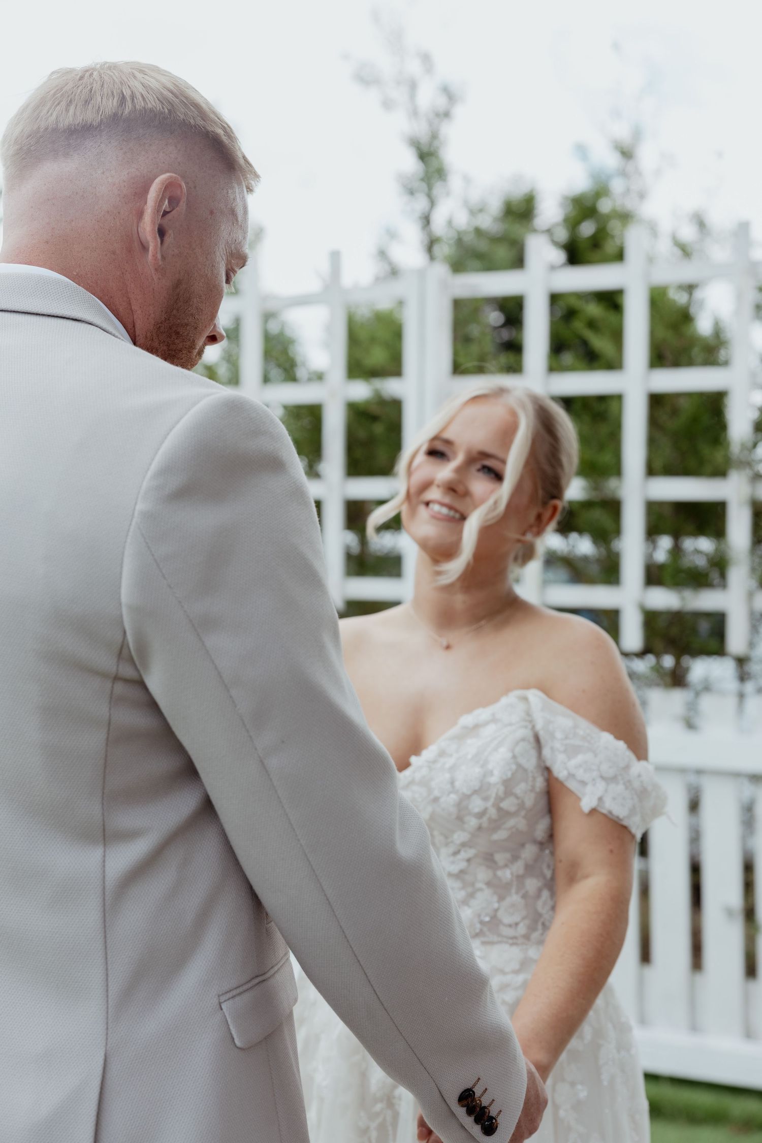 Bride in lace off-shoulder gown smiles at groom in gray suit during outdoor wedding ceremony.
