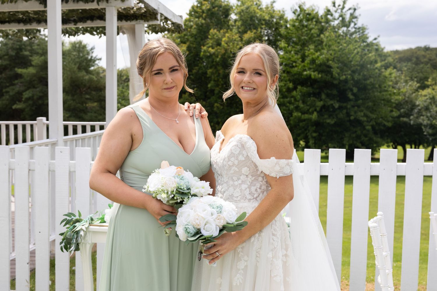 Bride in lace off-shoulder wedding gown posing with bridesmaid in sage green dress holding white bouquet.