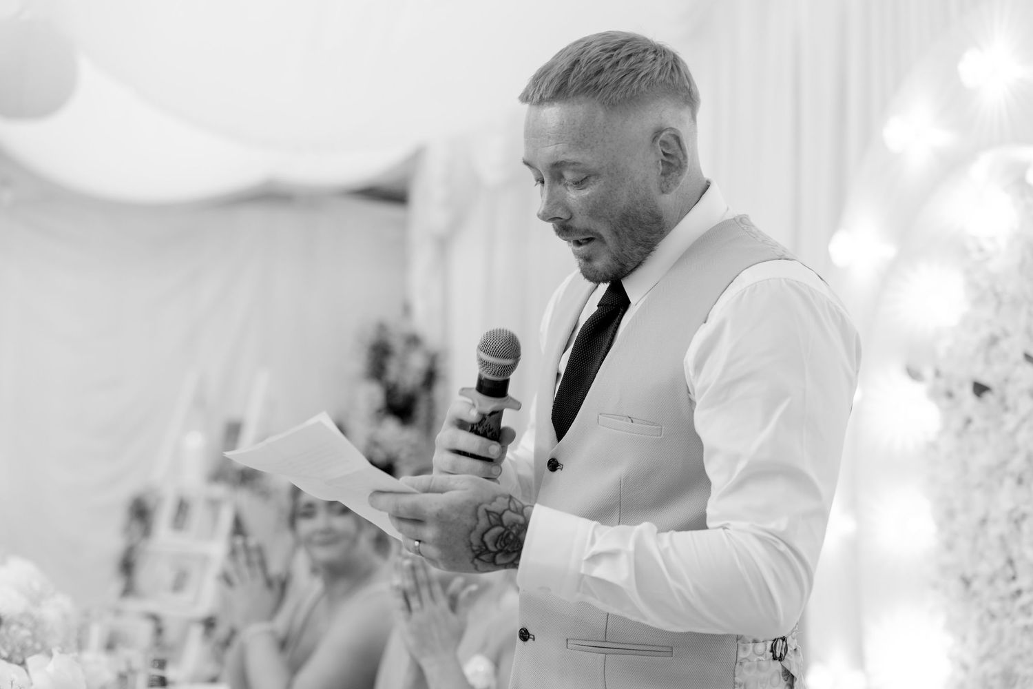 Groomsman reads emotional wedding speech into microphone at elegant black and white wedding reception.