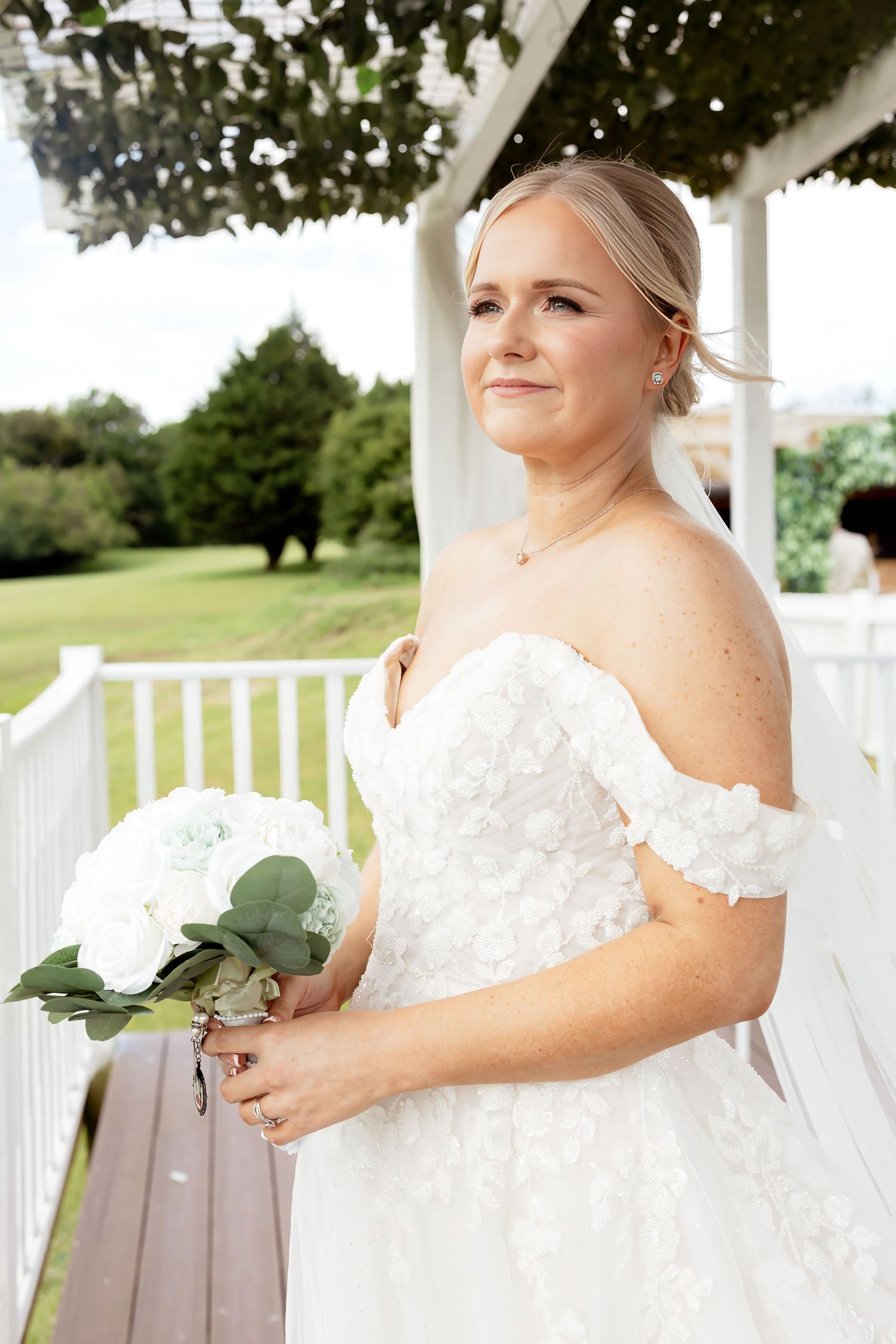 Beautiful bride in off-shoulder floral lace gown holding white hydrangea bouquet at pergola.