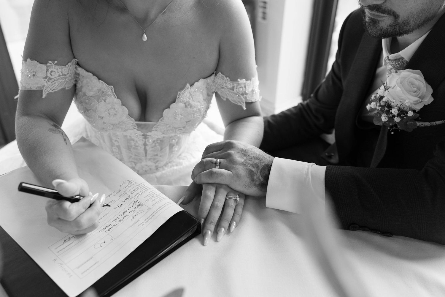 Black and white close-up of bride signing marriage register with groom placing hand on her arm.