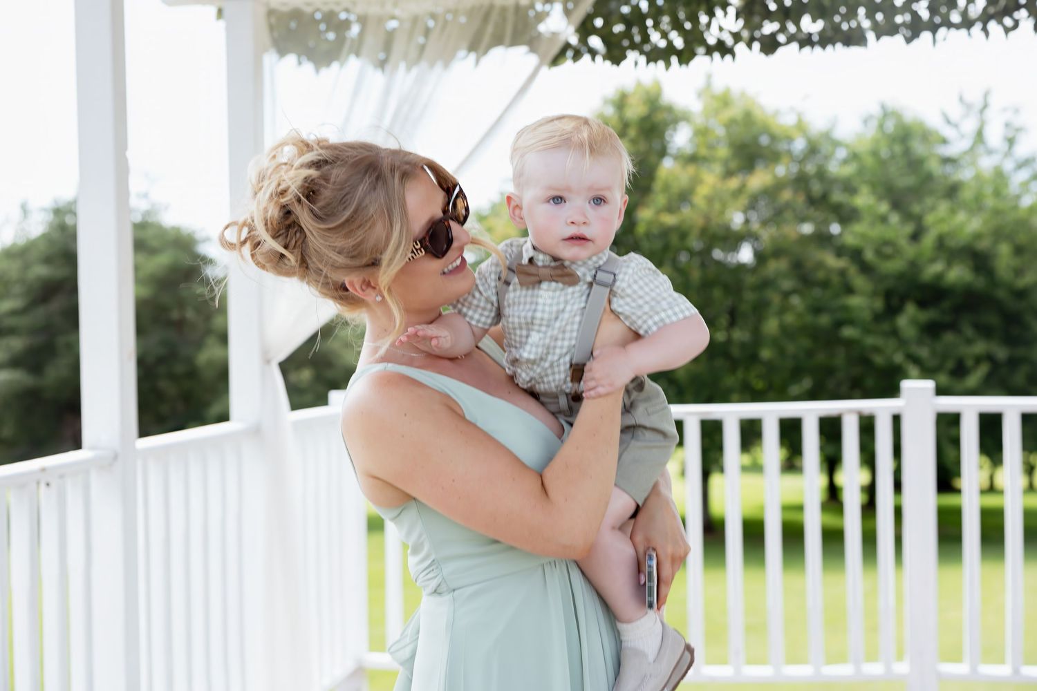 Woman in mint bridesmaid dress holding adorable baby boy in suspenders at outdoor wedding venue.