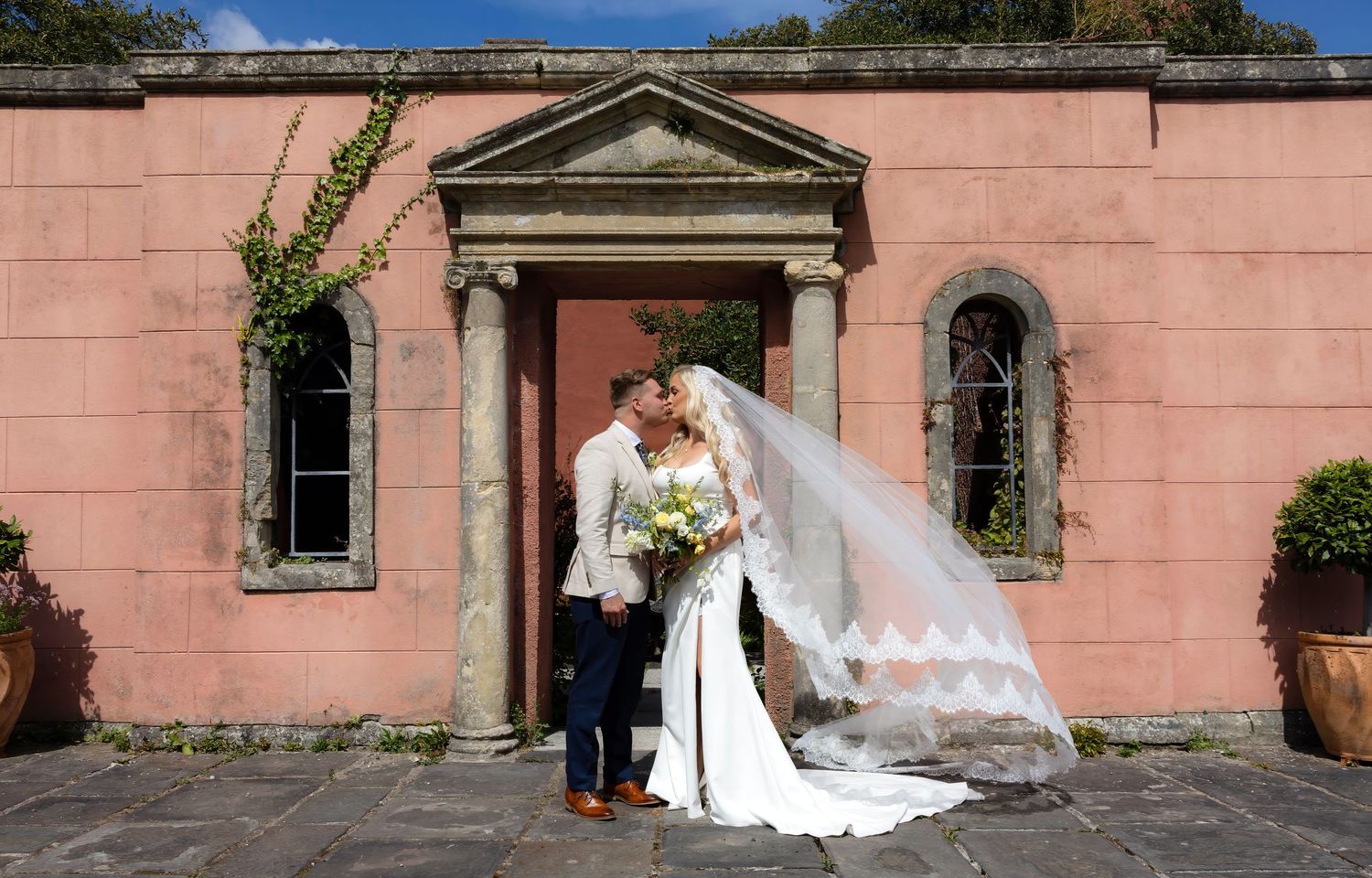 Bride and groom kissing outside a pink stone building, brides veil flowing in the wind, holding a white bouquet.