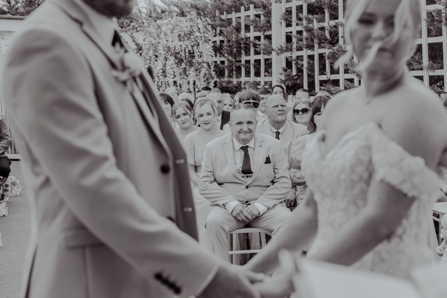 Black and white wedding ceremony photo showing bride and groom holding hands while guests watch emotionally in the background.