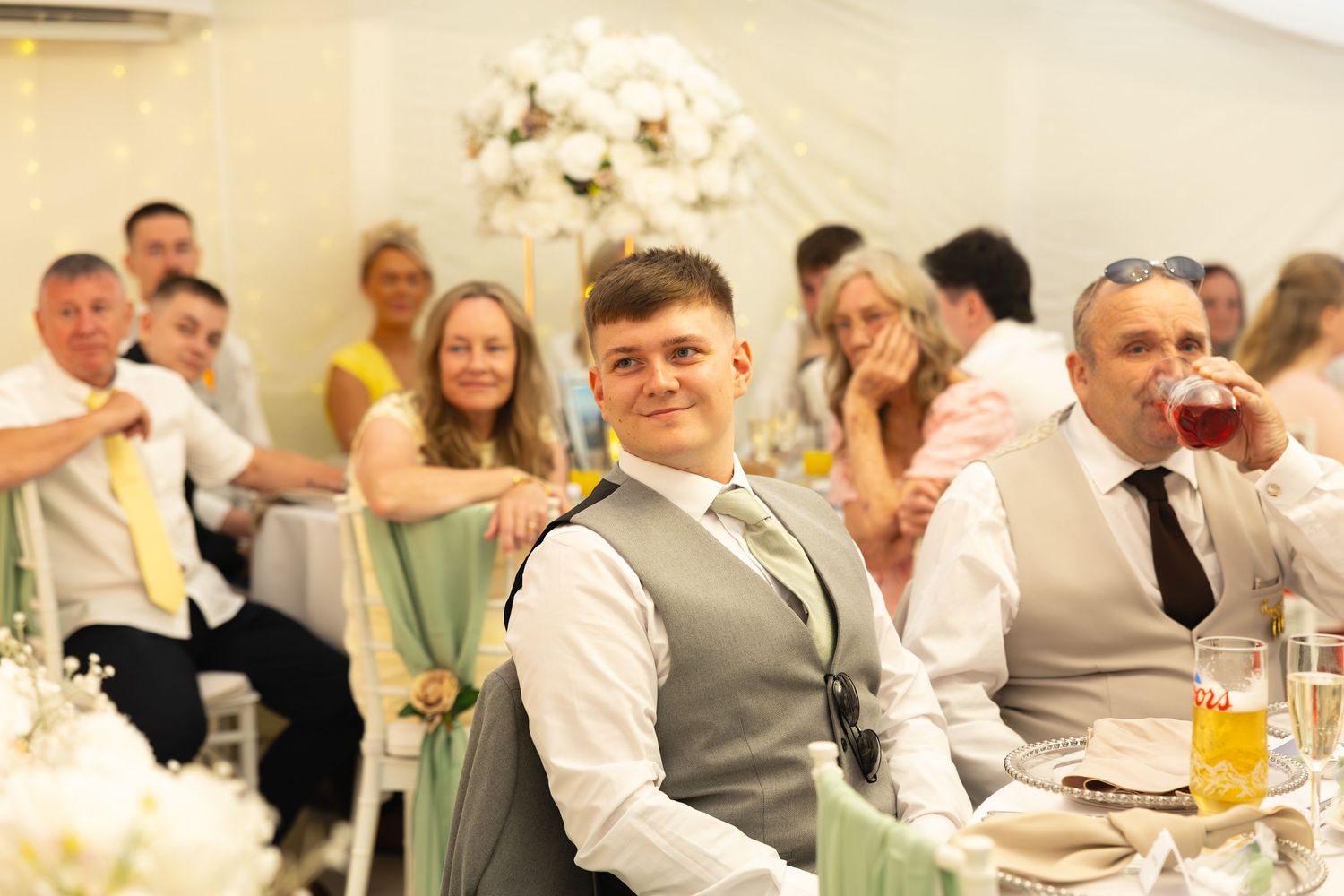 Young groomsman in grey waistcoat smiling at camera during elegant white floral wedding reception.