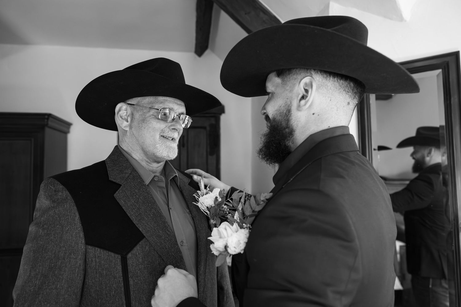 Two men in cowboy hats share a moment as one pins a boutonniere on the other in a black and white photo.