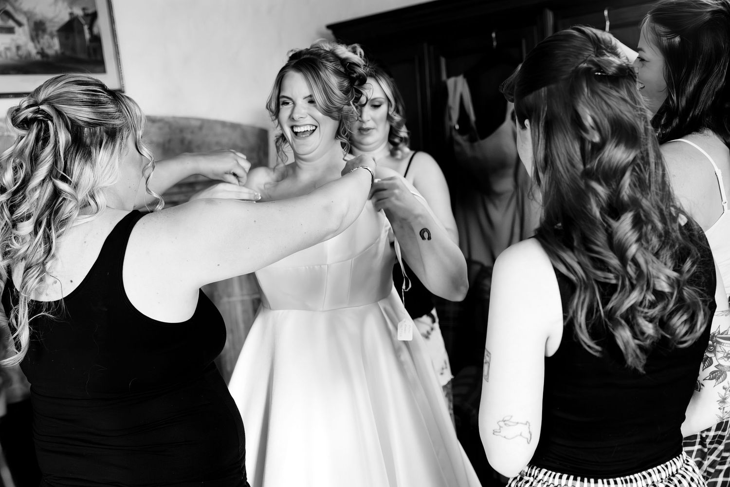 Bridesmaids helping a laughing bride into her wedding dress in a candid black and white getting ready photo.
