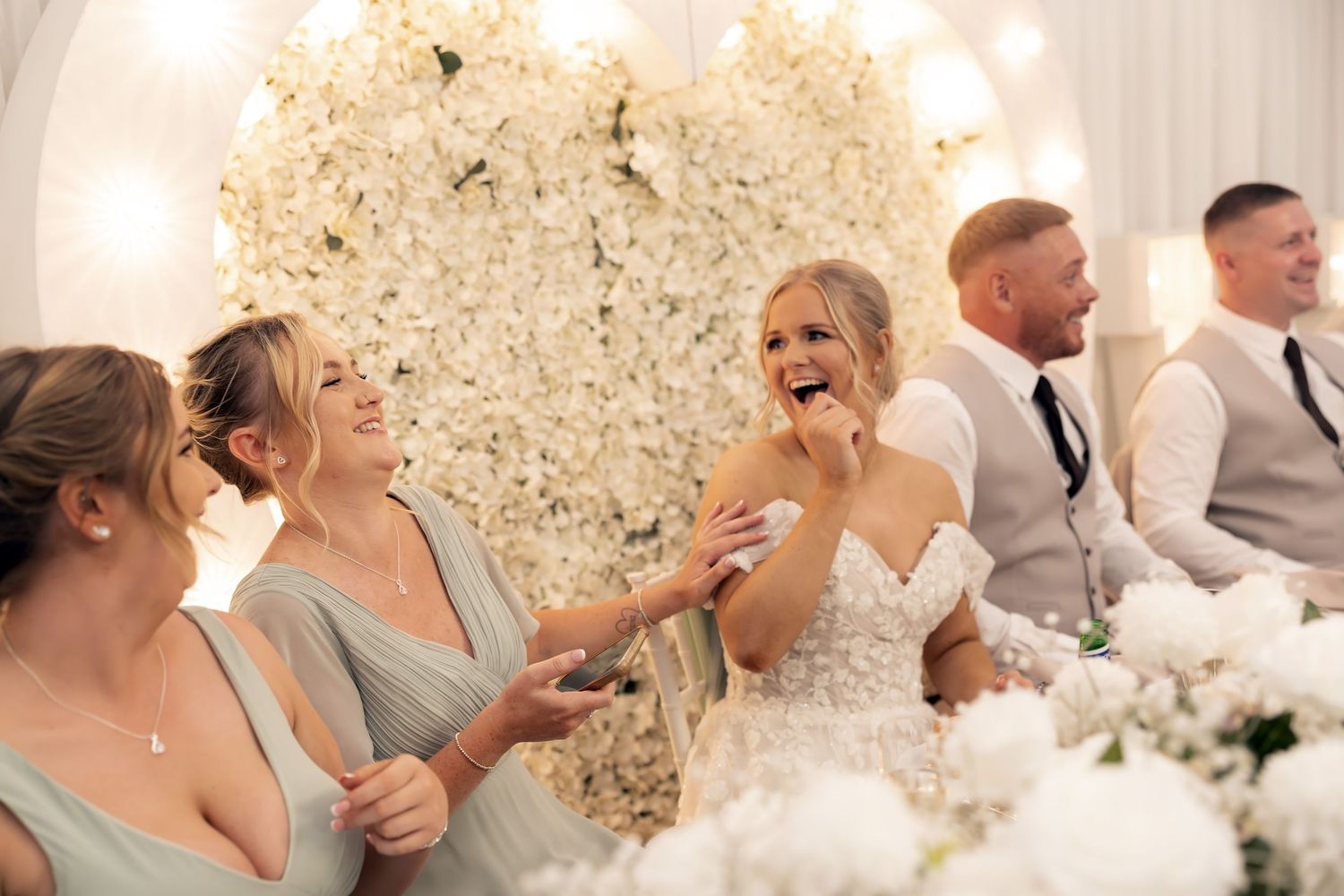 Bride and bridesmaids laugh joyfully at the wedding reception table adorned with white floral arrangements.