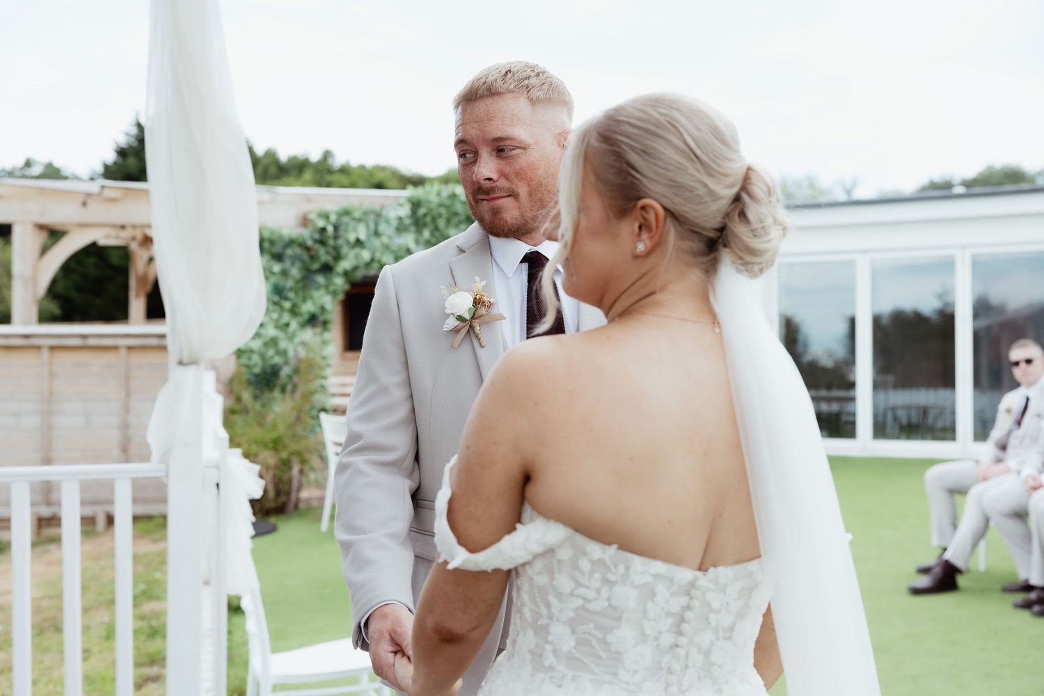 Bride and groom exchange vows outdoors at elegant wedding ceremony, groom in beige suit with boutonniere.