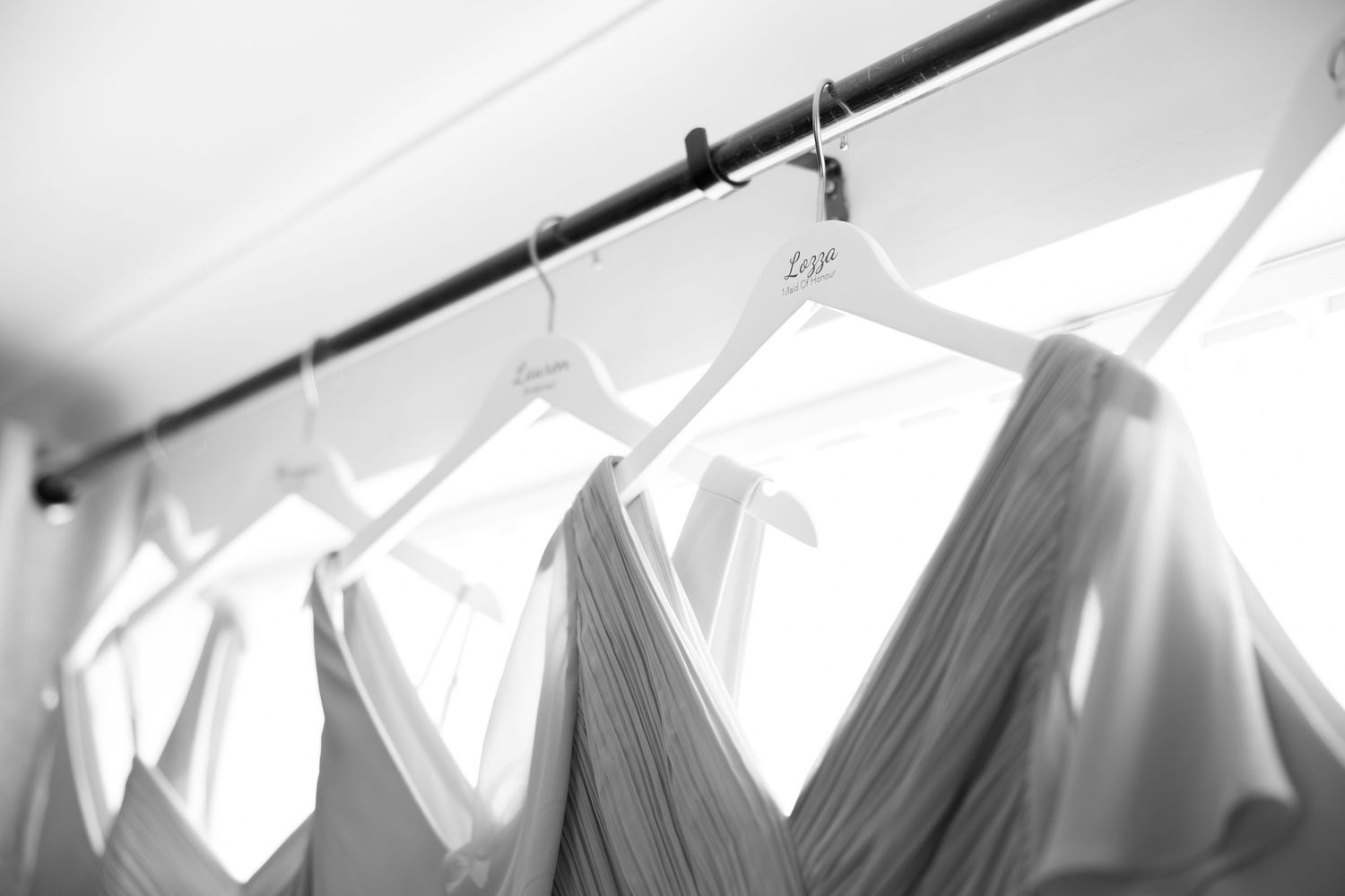 Black and white photo of bridesmaid dresses hanging on a rail with white hangers in a bright room.