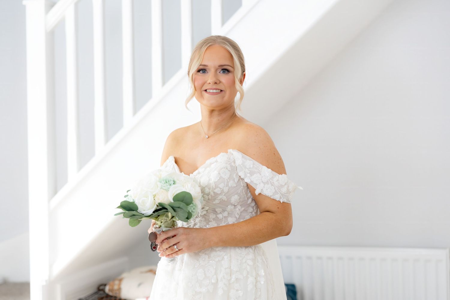 Smiling bride in off-shoulder lace wedding dress holding white bouquet near staircase.