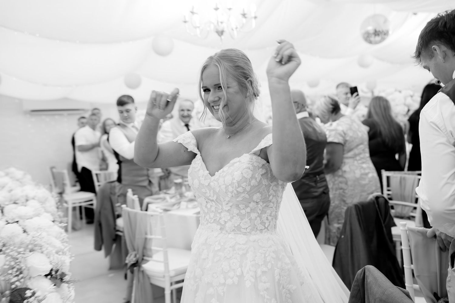 Joyful bride in lace wedding gown celebrates with raised fists in elegant reception hall with balloon decor.