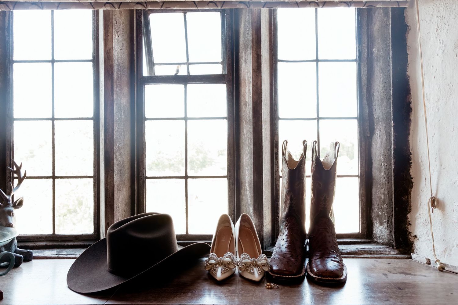 Wedding accessories including a black cowboy hat, white heels, and brown cowboy boots by a rustic window.