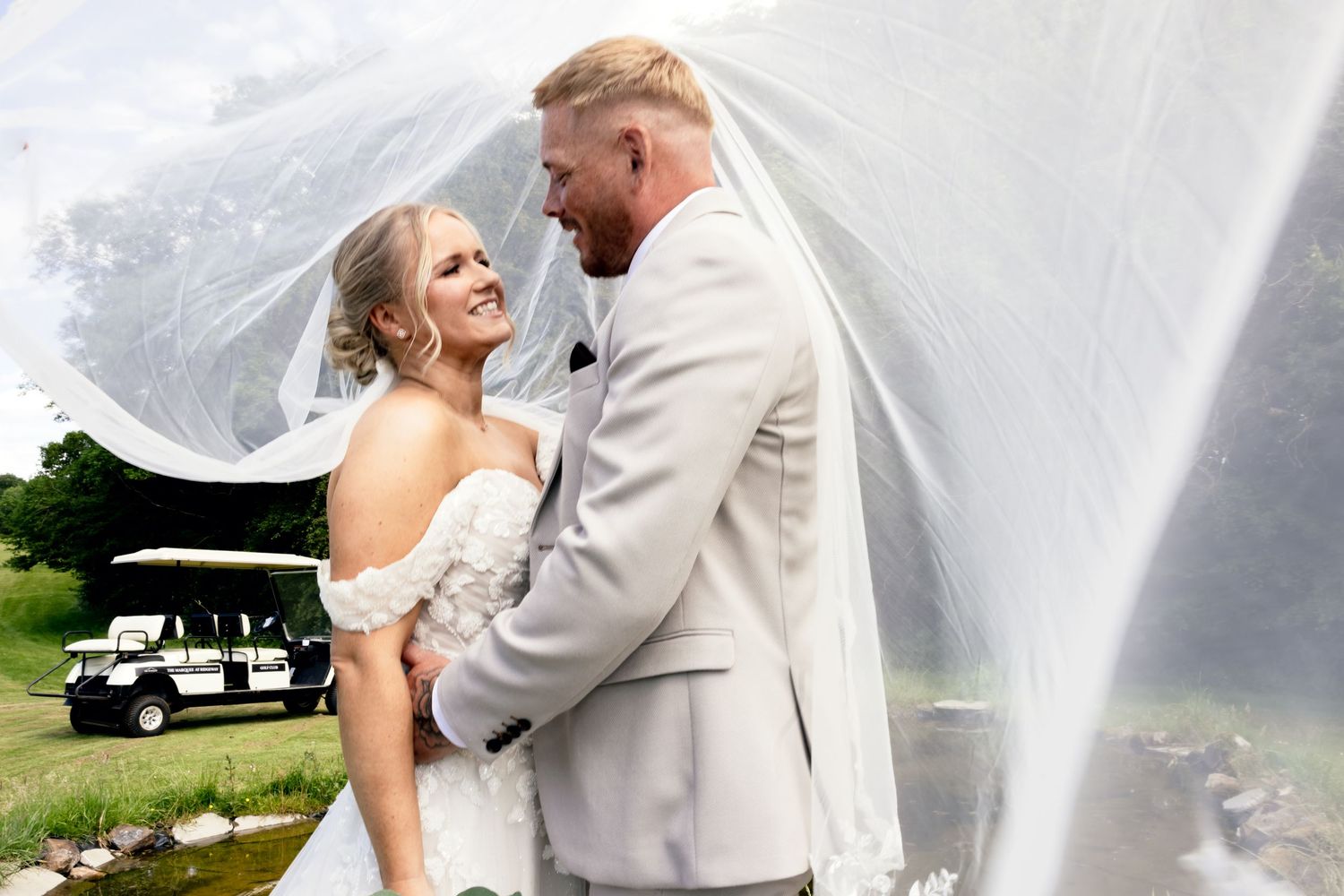 Bride and groom sharing a romantic moment under a flowing veil near a golf cart.