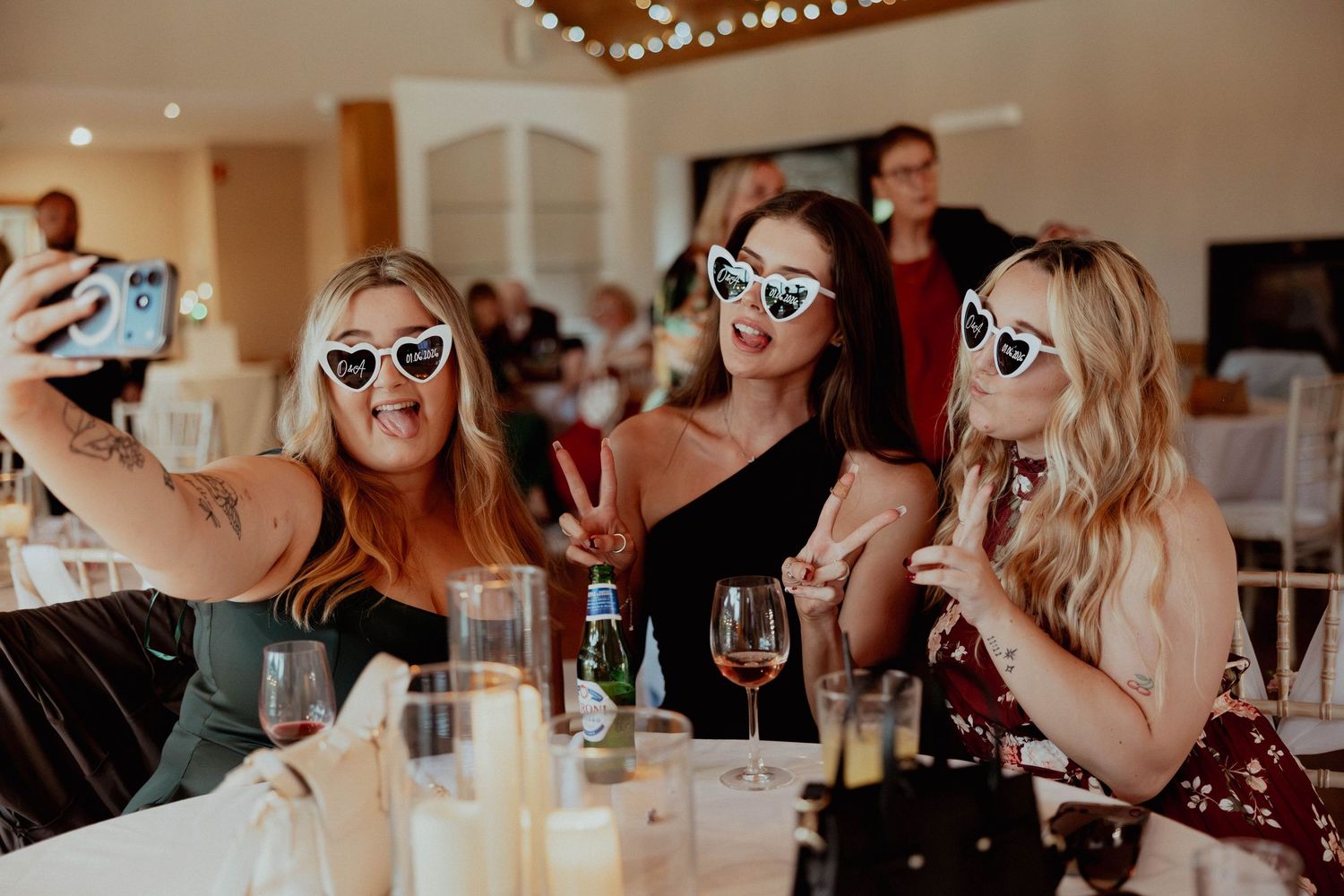 Three women wearing heart-shaped sunglasses taking a selfie at a wedding reception with wine glasses on the table.
