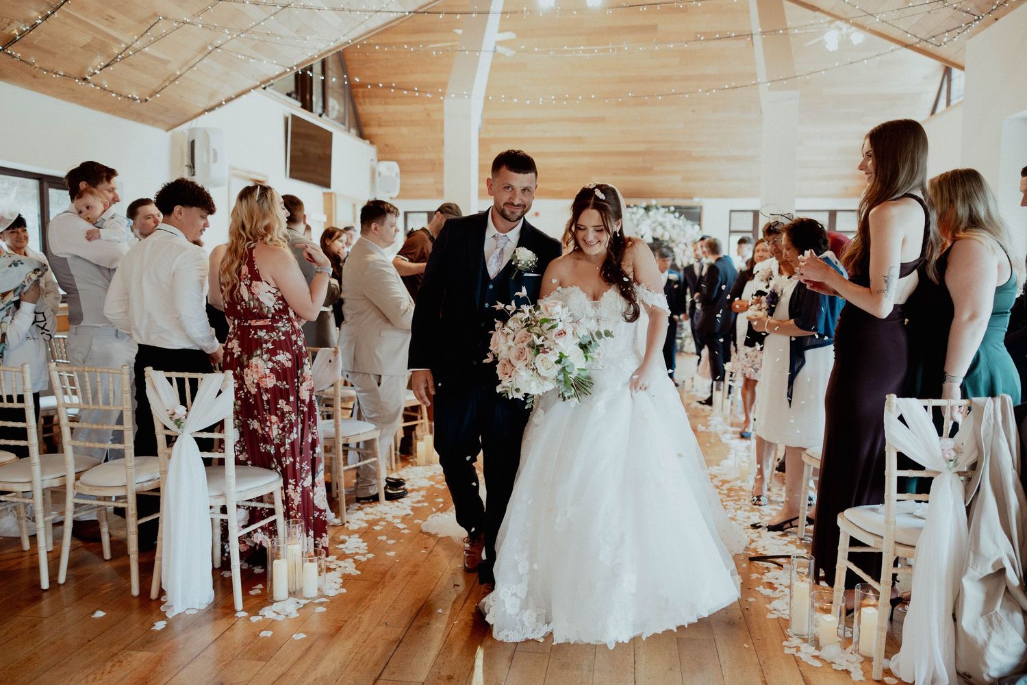 Bride and groom walk down the aisle together as guests celebrate with confetti at an elegant indoor wedding venue.