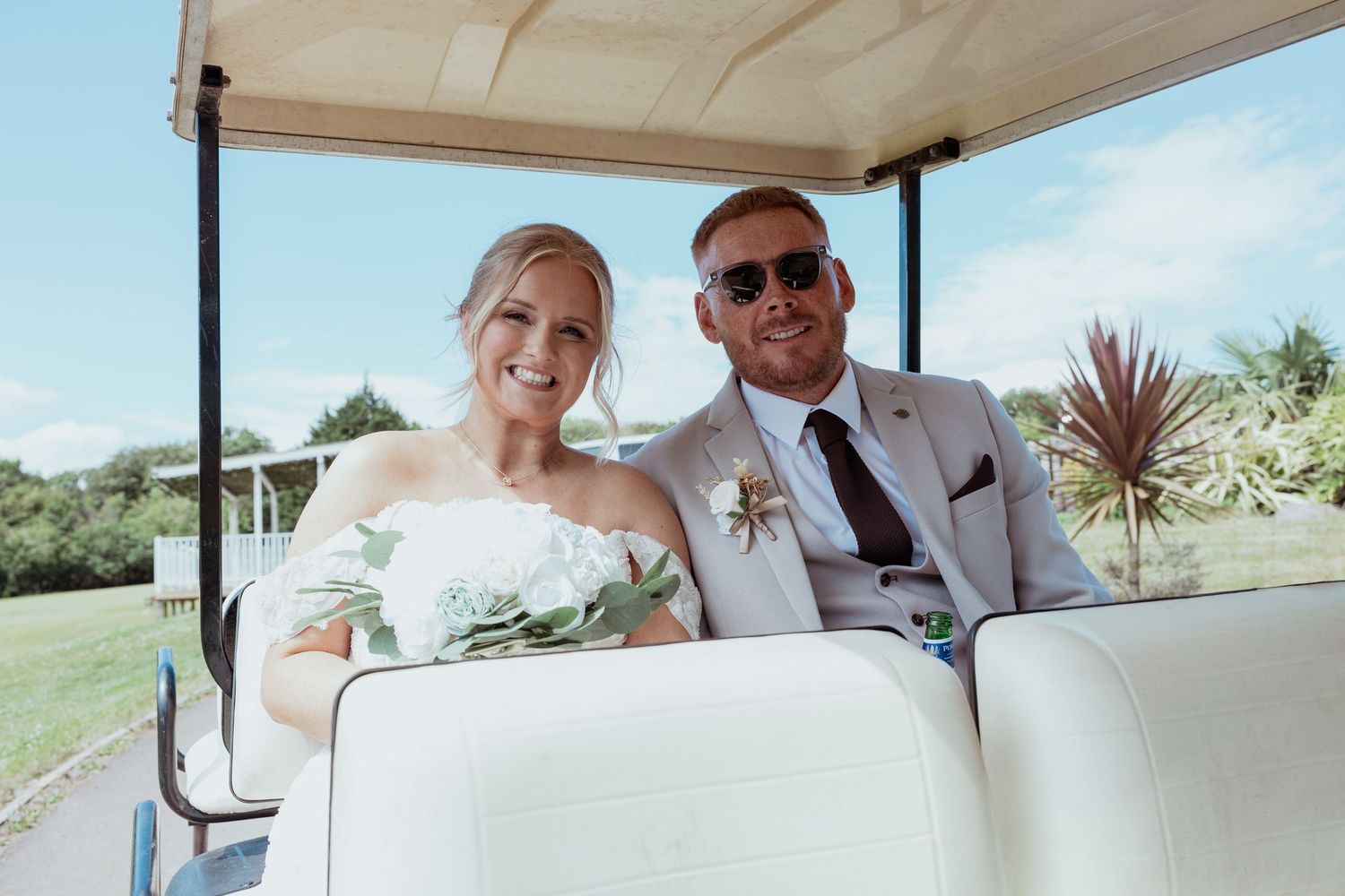 Smiling bride and groom riding golf cart in sunshine, bride holding white floral bouquet.