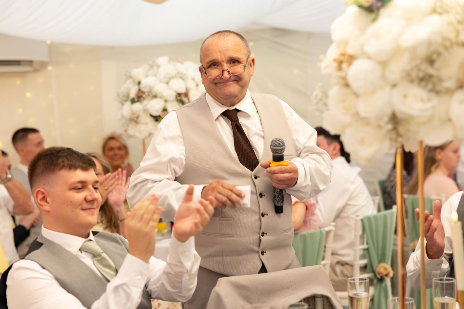Wedding guest delivers speech with microphone while guests applaud at white floral decorated reception.