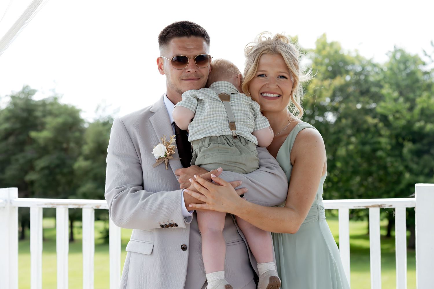 Smiling couple in wedding attire holding a baby in a checkered outfit at an outdoor wedding venue.