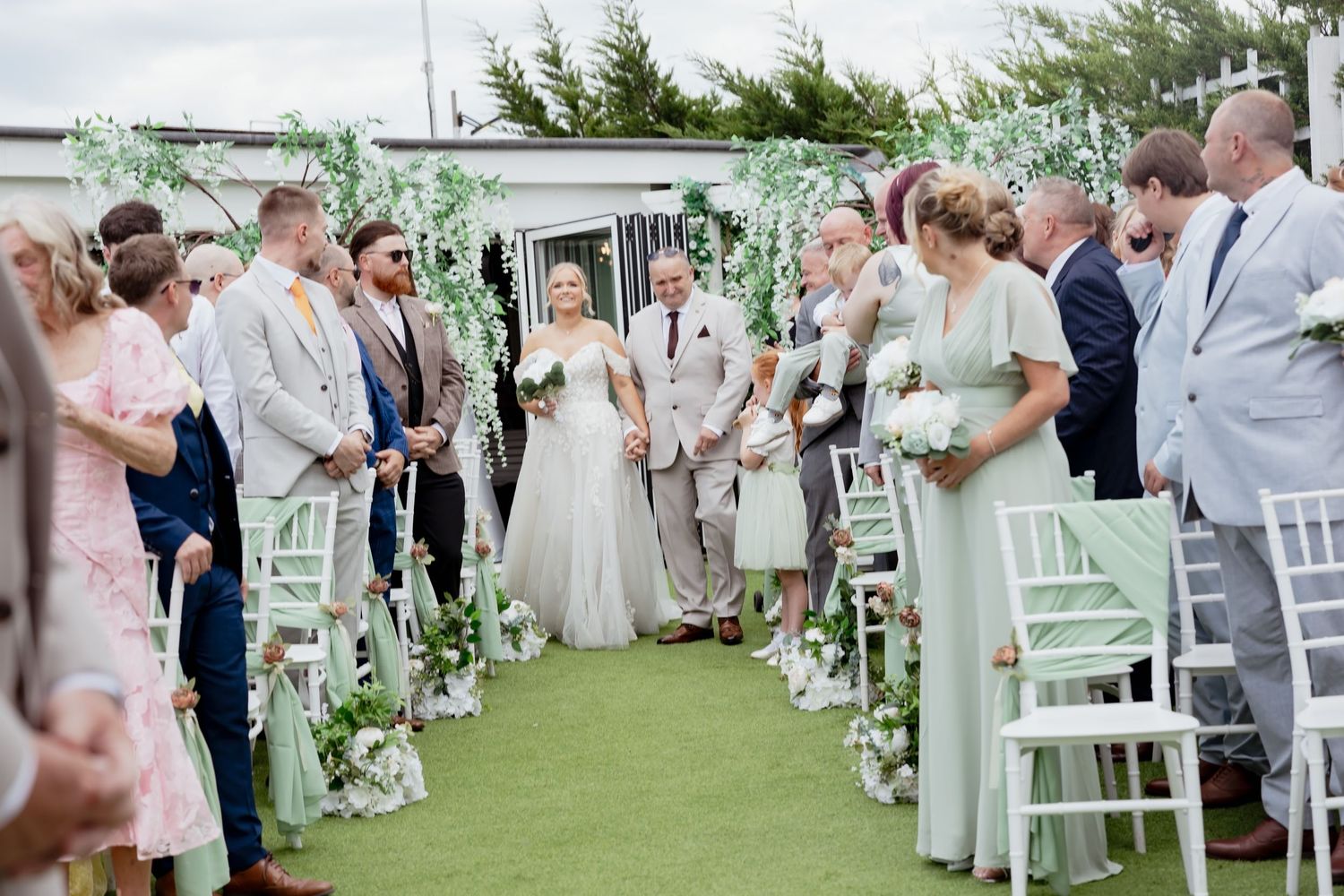 Bride walks down the aisle at an outdoor wedding ceremony with guests seated on white chairs.
