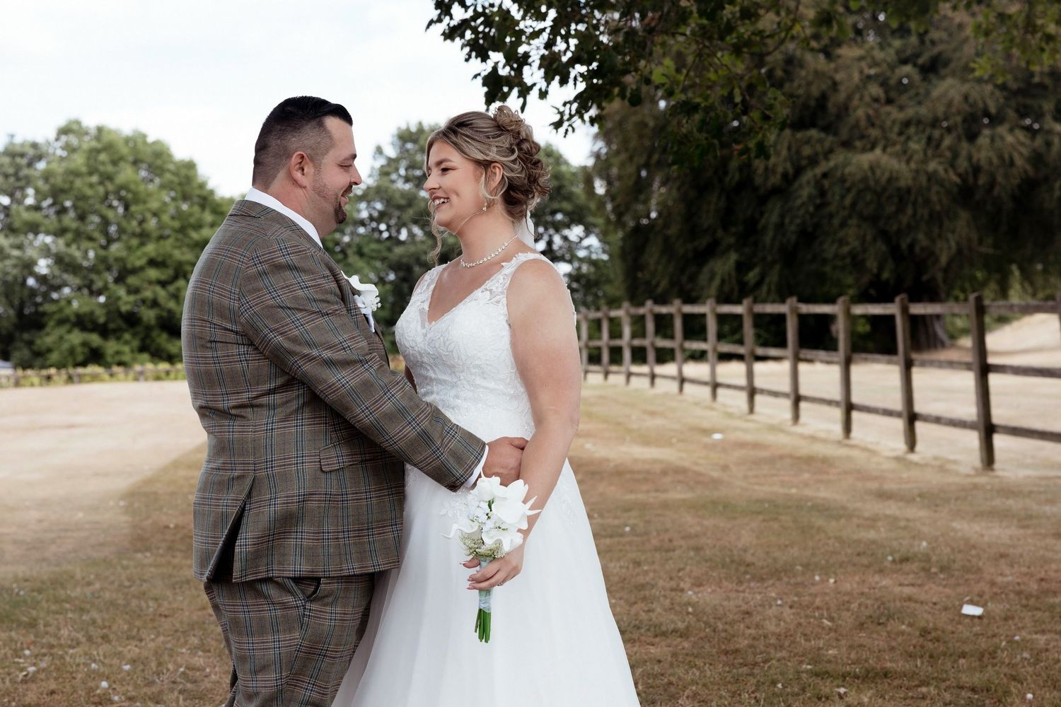 Bride holding white bouquet and groom embrace and smile at each other along a wooden fence outdoors.