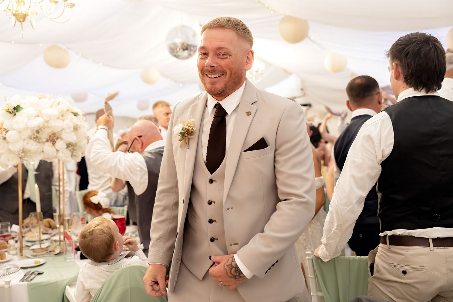 Smiling groom in beige suit and floral boutonniere at elegant wedding reception with white floral decor.