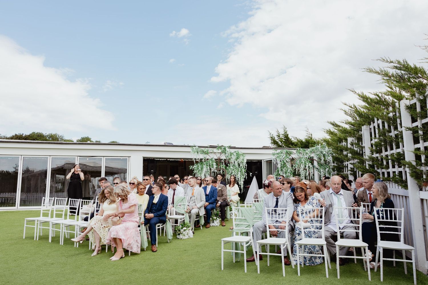 Outdoor wedding ceremony with guests seated on white chairs under blue sky.