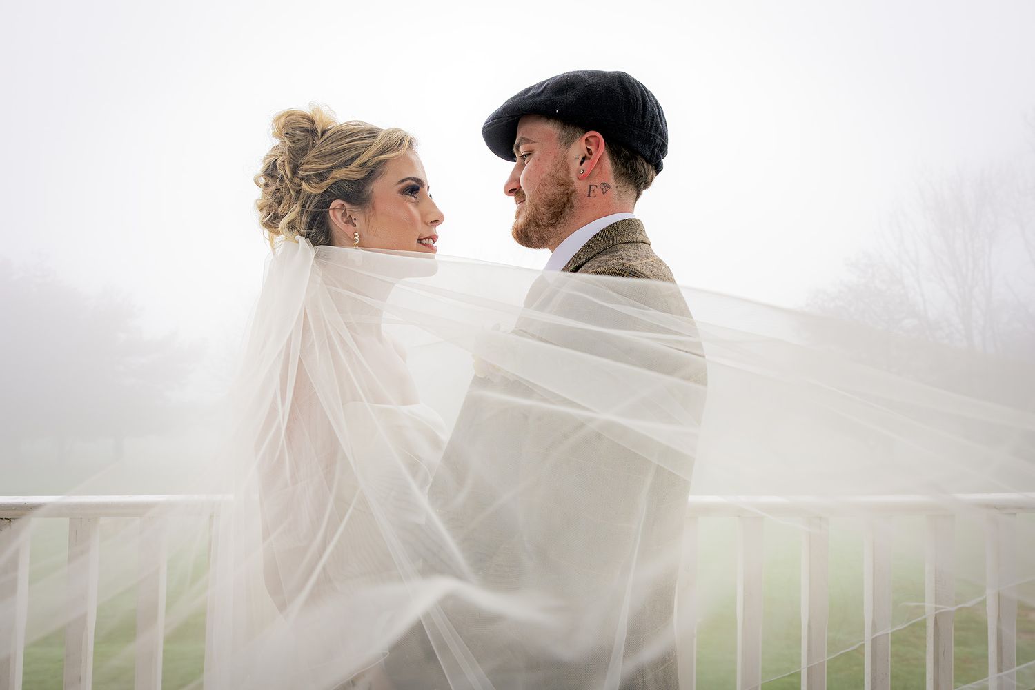 Bride and groom wrapped in flowing veil on foggy outdoor balcony, sharing romantic moment on wedding day. Ridgeway golf club, Caerphilly.