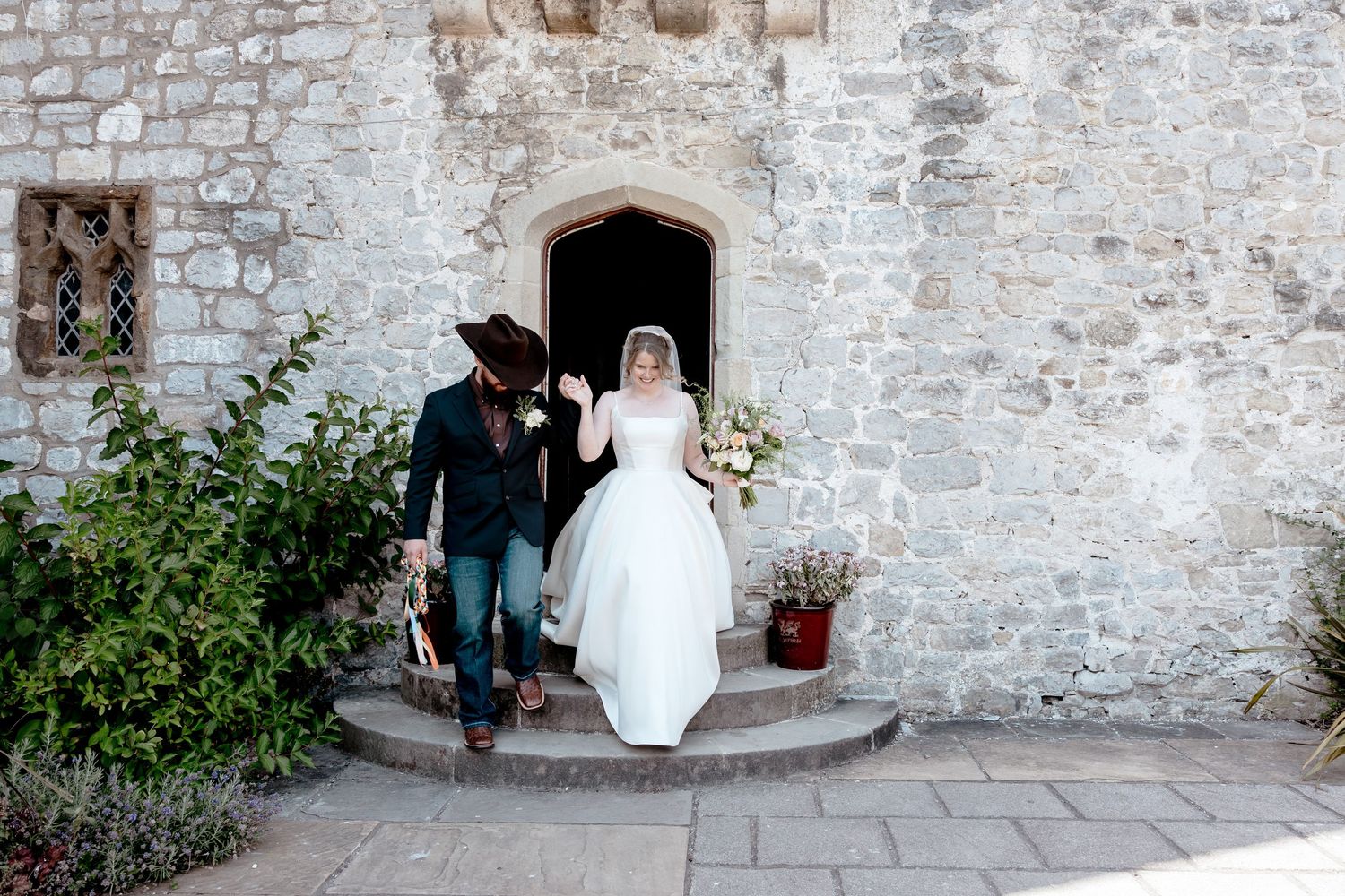Bride and groom exit a rustic stone church, groom wearing a cowboy hat and jeans on their wedding day.
