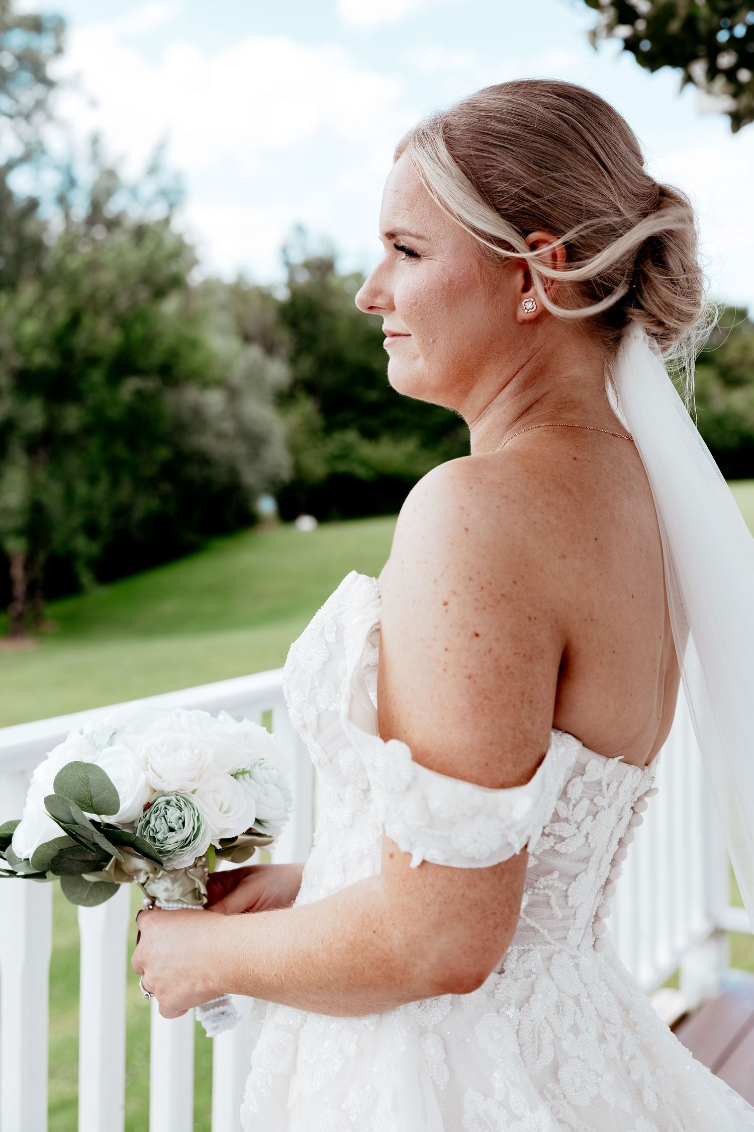 Elegant bride in off-shoulder lace gown holding white bouquet on a white railed balcony outdoors.