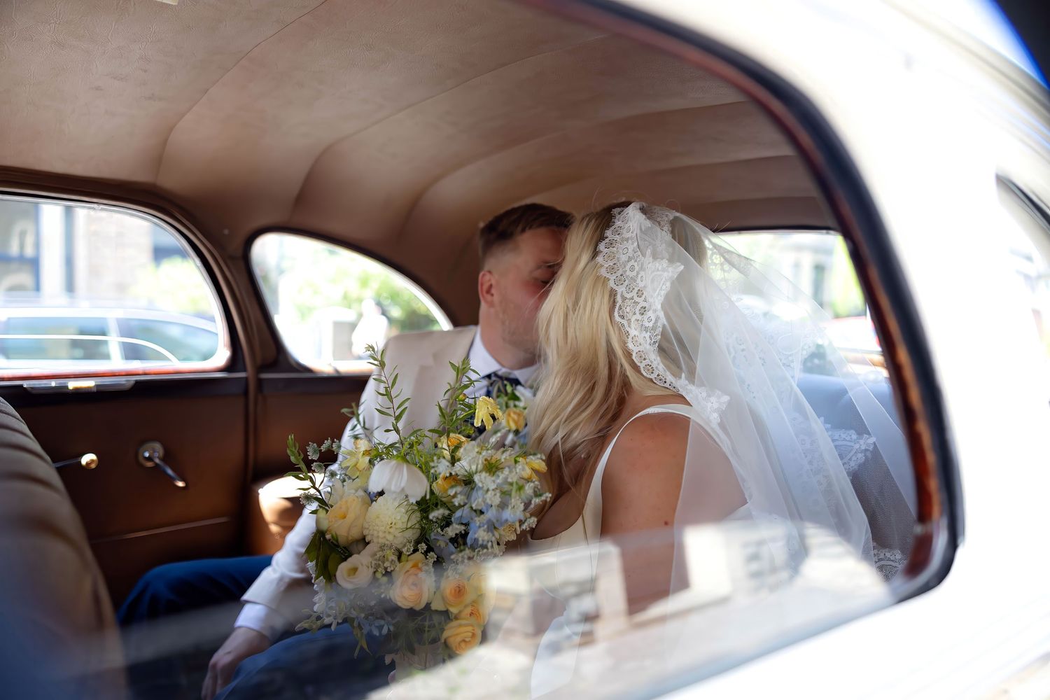 Bride and groom kissing in vintage wedding car, bride holding yellow and white bouquet wearing veil.