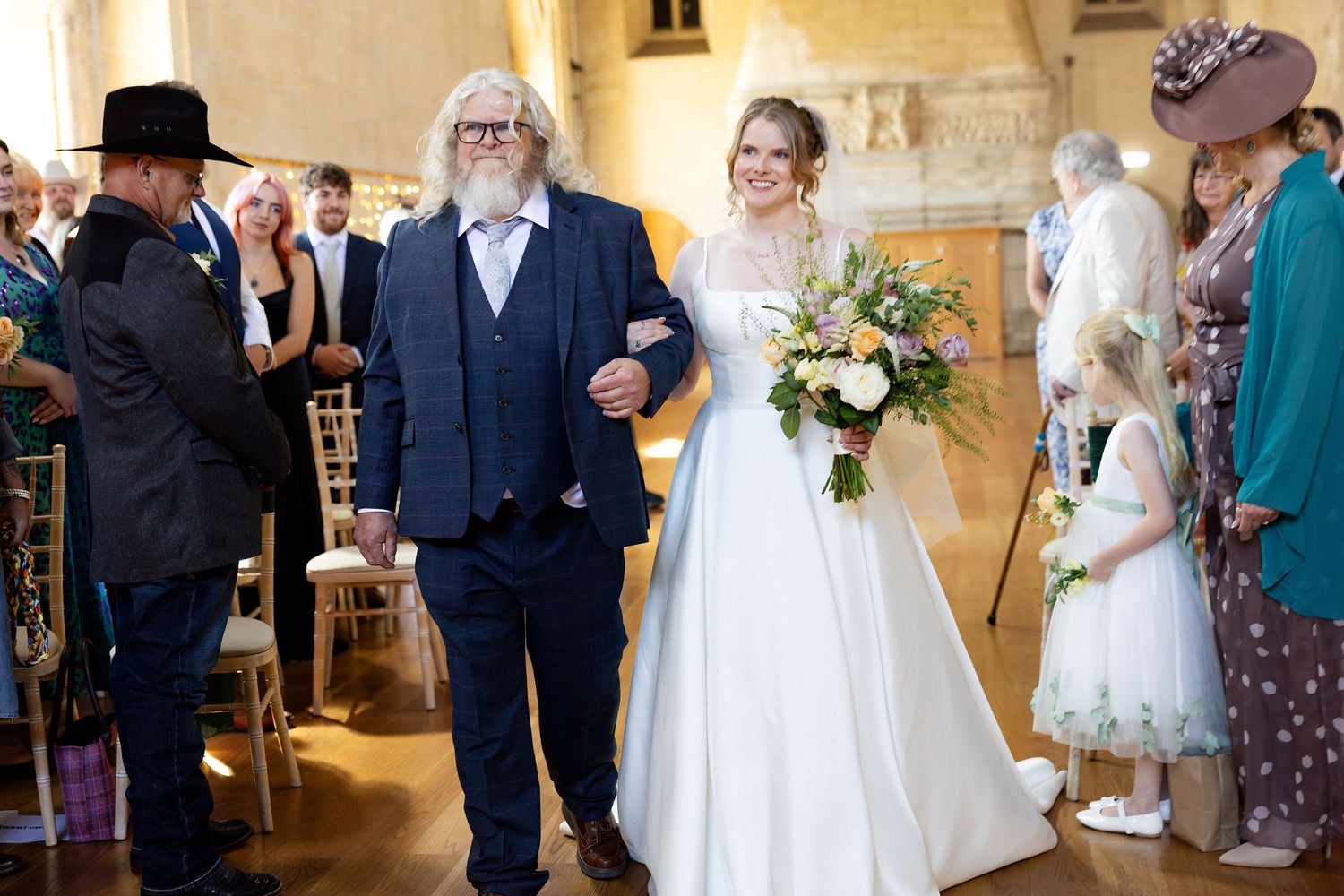 Father walks bride down the aisle in a historic stone chapel, bride carrying a colorful spring bouquet.